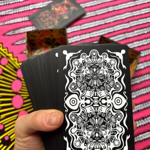 May include: A deck of black tarot cards with a white floral design, held in a hand. The cards are fanned out, revealing the intricate pattern. A box with the same design is visible in the background, along with a pink and white striped fabric.