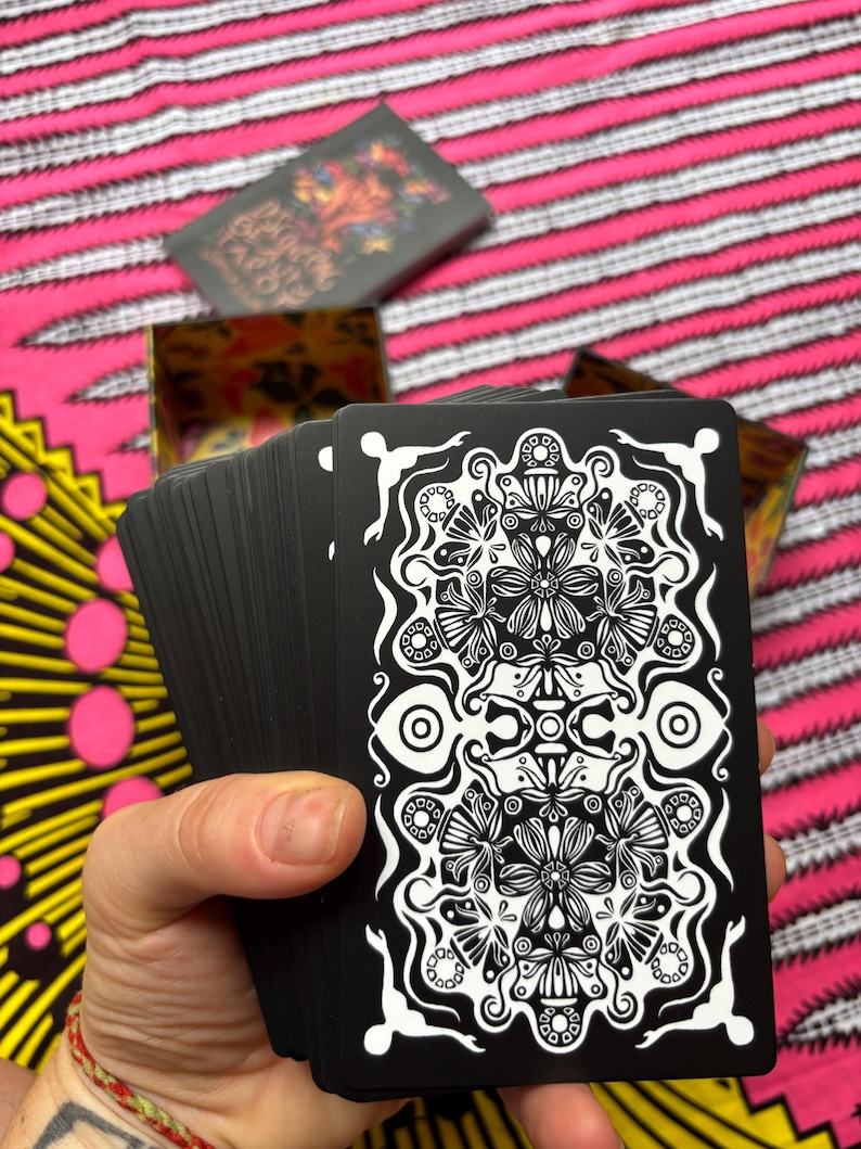 May include: A deck of black tarot cards with a white floral design, held in a hand. The cards are fanned out, revealing the intricate pattern. A box with the same design is visible in the background, along with a pink and white striped fabric.