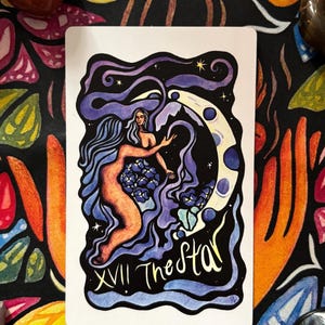May include: A tarot card featuring an illustration of a nude woman with long blue hair reaching towards a crescent moon. The card is titled "The Star" with the Roman numeral XVII. The background is a dark floral pattern.