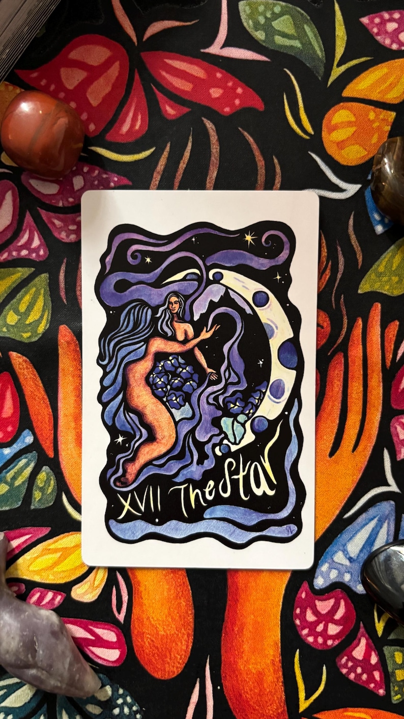 May include: A tarot card featuring an illustration of a nude woman with long blue hair reaching towards a crescent moon. The card is titled "The Star" with the Roman numeral XVII. The background is a dark floral pattern.