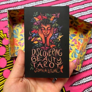 May include: A black tarot card featuring a woman with butterflies in her hair and the text "Discovering Beauty Tarot" by Sophia Litwak. The card is held in a hand, with a colorful box in the background.