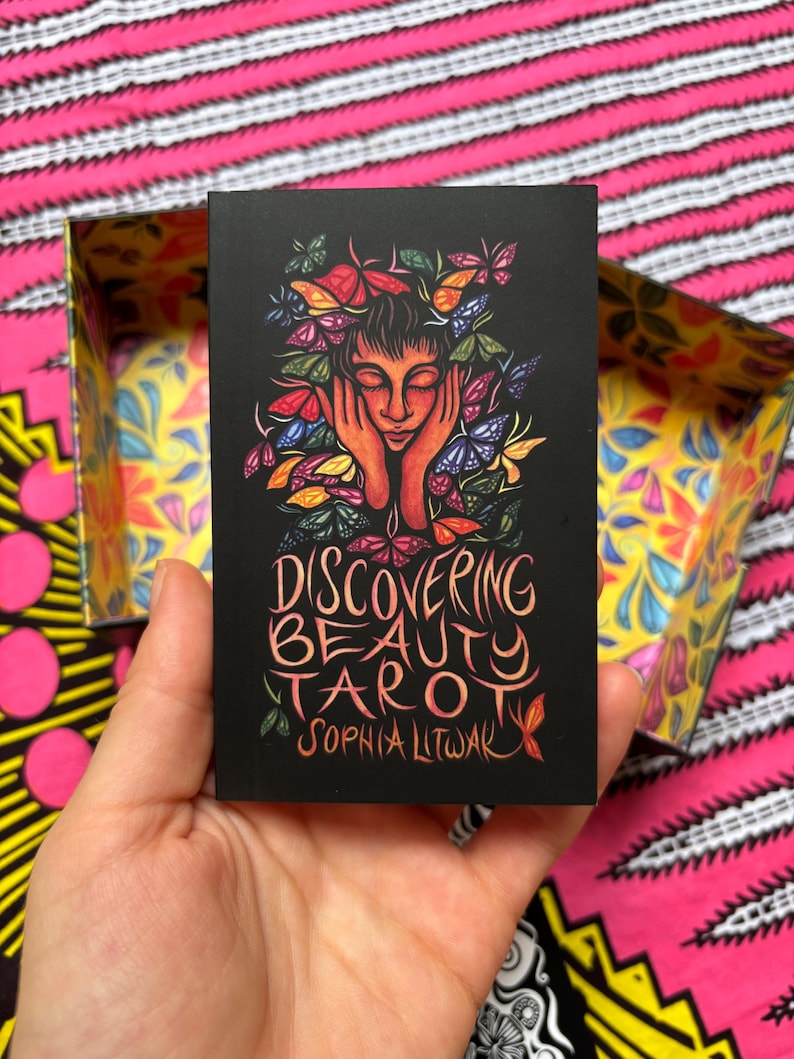 May include: A black tarot card featuring a woman with butterflies in her hair and the text "Discovering Beauty Tarot" by Sophia Litwak. The card is held in a hand, with a colorful box in the background.