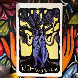 May include: A tarot card featuring two figures under a tree with a yellow background. The card is labeled "XI Justice" in black script. The background is a black cloth with colorful floral and hand designs.