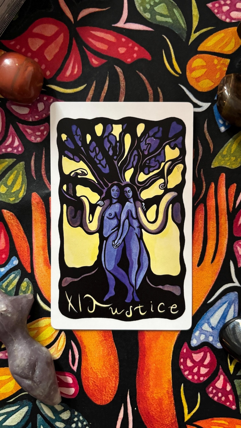 May include: A tarot card featuring two figures under a tree with a yellow background. The card is labeled "XI Justice" in black script. The background is a black cloth with colorful floral and hand designs.