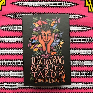 May include: A black tarot card box with the title "Discovering Beauty Tarot" and the author's name, Sophia Litwak, in orange script. The box features an illustration of a person with butterflies around their head. The background is a pink and white patterned fabric.
