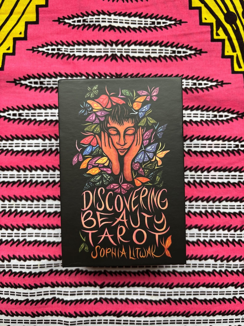 May include: A black tarot card box with the title "Discovering Beauty Tarot" and the author's name, Sophia Litwak, in orange script. The box features an illustration of a person with butterflies around their head. The background is a pink and white patterned fabric.