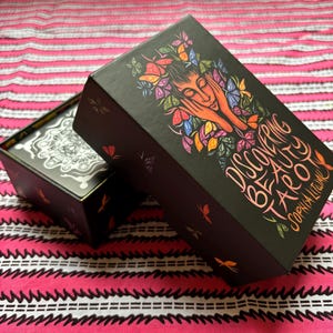 May include: Black tarot card box with the title "Discovering Beauty Tarot" and the author's name. The box features a colorful illustration of a woman's face surrounded by butterflies and flowers. The box is open, revealing the cards inside.