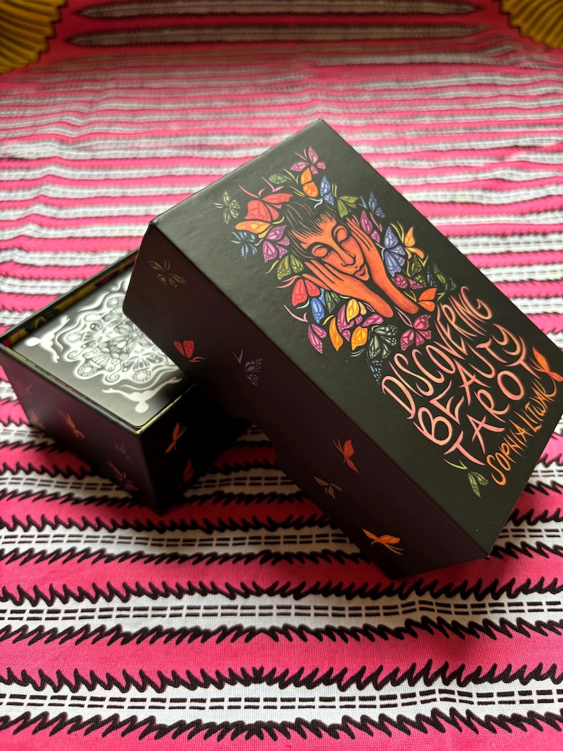 May include: Black tarot card box with the title "Discovering Beauty Tarot" and the author's name. The box features a colorful illustration of a woman's face surrounded by butterflies and flowers. The box is open, revealing the cards inside.
