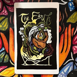 May include: A tarot card with the words "The Earth" in script. The card features a colorful illustration of a figure with a flame, surrounded by a snake, bird, and other elements, set against a black background. The card is on a floral patterned surface.