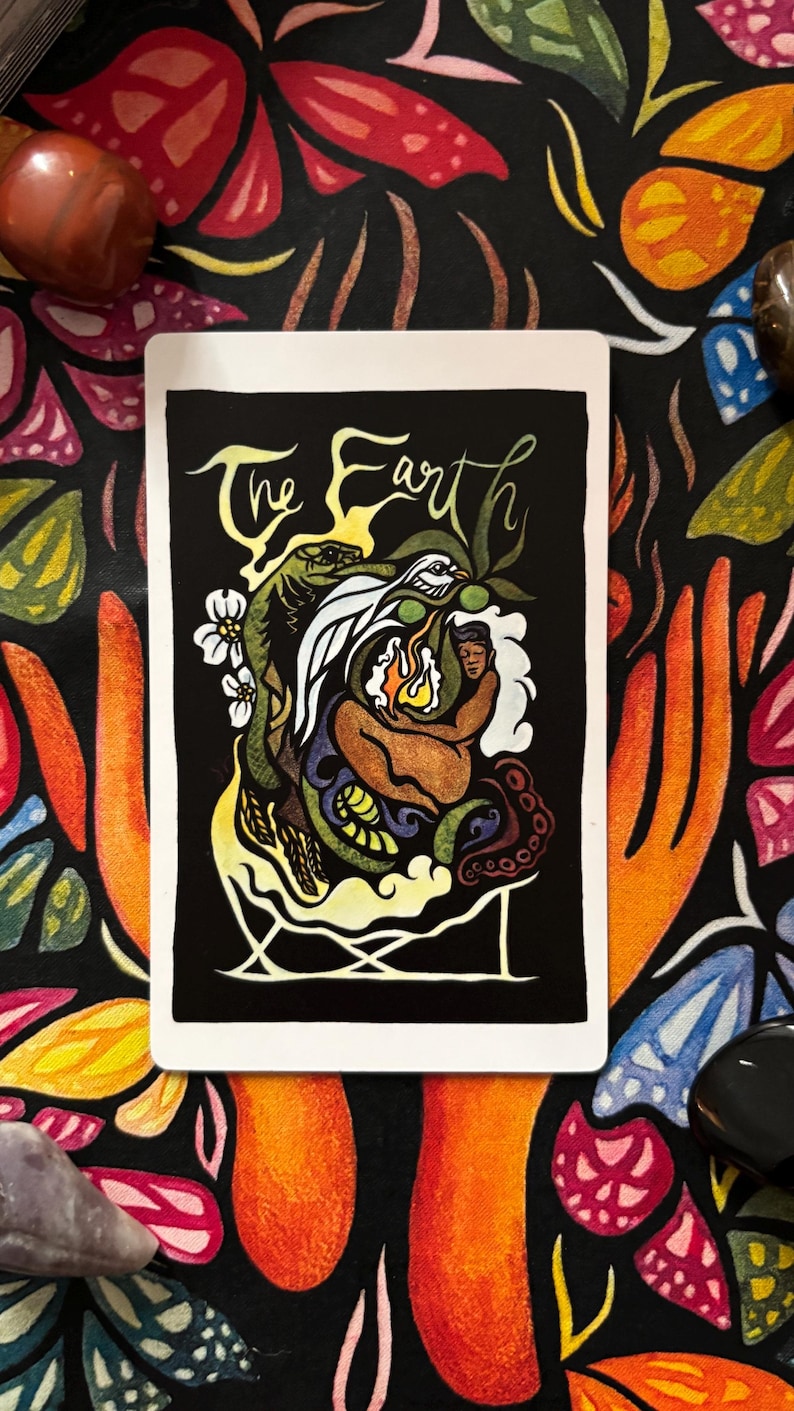 May include: A tarot card with the words "The Earth" in script. The card features a colorful illustration of a figure with a flame, surrounded by a snake, bird, and other elements, set against a black background. The card is on a floral patterned surface.