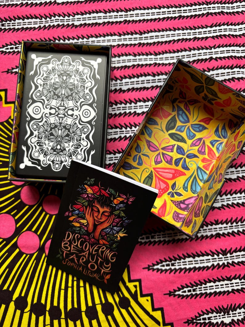 May include: A tarot card set with a black box featuring a white floral design and a yellow box with a butterfly pattern. The set includes a black booklet titled "Discovering Beauty Tarot" by Sophia Litwak.