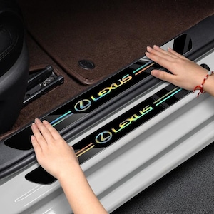 May include: Black car door sill protectors with the Lexus logo in iridescent colors. The protectors have a sleek, modern design and are being installed in a vehicle. The Lexus logo is centered with a rainbow-colored stripe.