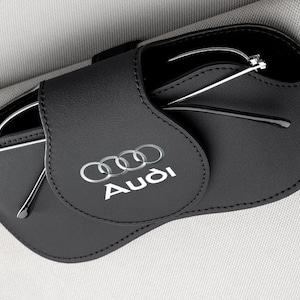 May include: Black leather eyeglass case with the Audi logo in silver. The case is open, revealing a pair of silver-framed glasses. The case is designed to hold and protect eyeglasses.