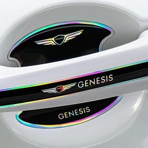 May include: Car door handle accents with the Genesis logo and text. The black accents have a rainbow iridescent trim. The Genesis logo is a winged emblem with a red center. The car door is white.