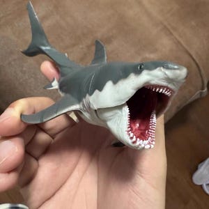 Pode incluir: Um brinquedo de tubarão cinza e branco com a boca aberta revelando fileiras de dentes brancos e um interior vermelho. O tubarão tem barbatanas e é segurado em uma mão. O brinquedo é uma representação detalhada de um tubarão branco.