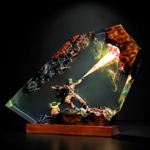 May include: A resin sculpture depicting a Mandalorian figure firing a blaster, with Grogu beside him. The figures are encased in a clear, angled resin block with colourful, textured details and a wooden base.