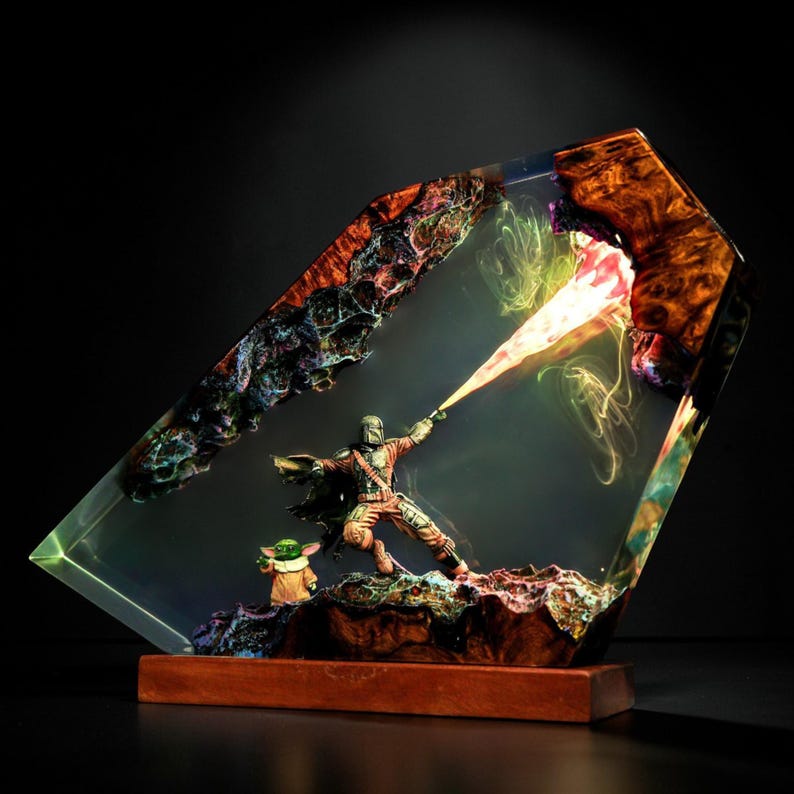 May include: A resin sculpture depicting a Mandalorian figure firing a blaster, with Grogu beside him. The figures are encased in a clear, angled resin block with colourful, textured details and a wooden base.