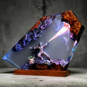 May include: A resin sculpture of a Mandalorian figure with Grogu, set within a clear, angled resin block. The sculpture has a wooden base and features a blaster effect. The resin block has a mix of colours, including purple, blue, and orange.