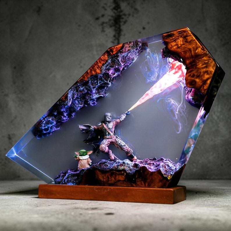 May include: A resin sculpture of a Mandalorian figure with Grogu, set within a clear, angled resin block. The sculpture has a wooden base and features a blaster effect. The resin block has a mix of colours, including purple, blue, and orange.