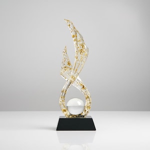 May include: An abstract clear sculpture with gold leaf detailing, featuring a swirling design and a spherical element. The sculpture is mounted on a black rectangular base, creating a modern and elegant aesthetic.