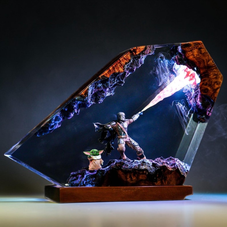 May include: A Star Wars-themed resin art piece featuring The Mandalorian and Grogu. The artwork is encased in a clear, angled resin block with a wooden base. The Mandalorian is depicted with a blaster, and Grogu is also present.
