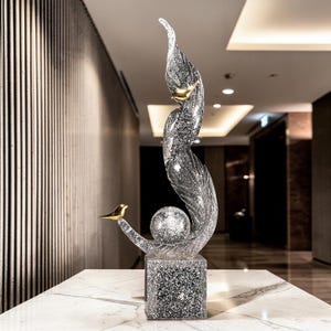 May include: A decorative glass sculpture featuring a swirling, feather-like design with a gold-colored bird perched on a curved branch. The sculpture includes a sphere and sits on a square base with a speckled pattern. The artwork is displayed on a marble surface.