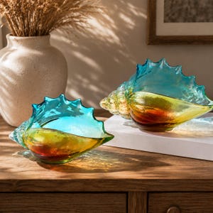 May include: Two decorative glass conch shells featuring a gradient color transition from deep amber at the base to a vibrant turquoise blue at the top edge. The shells have a translucent, smooth finish with a spiraled structure and serrated edges.