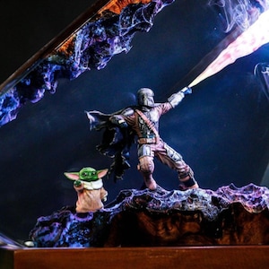 May include: A detailed diorama featuring The Mandalorian figure with a blaster beam, and Grogu. The figures are set against a dark blue backdrop with purple and blue accents. The base is brown and textured.