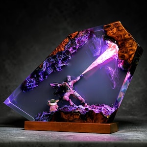 May include: A resin sculpture featuring a Mandalorian figure with a purple energy beam, and a small Grogu figure. The sculpture is encased in a clear resin with purple and orange accents, mounted on a wooden base.