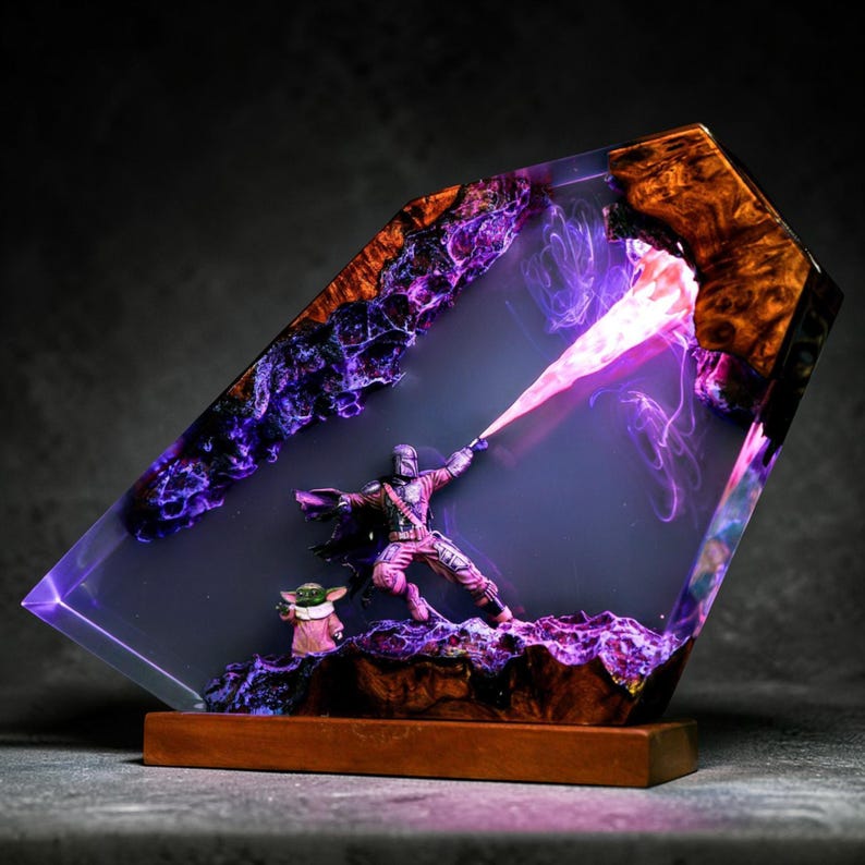May include: A resin sculpture featuring a Mandalorian figure with a purple energy beam, and a small Grogu figure. The sculpture is encased in a clear resin with purple and orange accents, mounted on a wooden base.