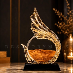 May include: A clear, curved glass sculpture containing golden flakes and a mound of brown sand, set within a circular frame. The piece is mounted on a solid black rectangular base. The text DESIGNBYMLS is visible in the background.