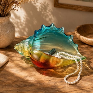 Può includere: Una ciotola decorativa in vetro a forma di conchiglia con una vivace sfumatura di colori che passa dall'ambra profondo alla base al giallo al centro e al turchese brillante sul bordo superiore, contenente un orologio da polso e una collana di perle.