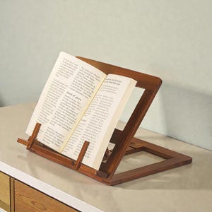May include: A wooden book stand with an open book. The stand is made of dark brown wood and has a slanted design to hold the book upright. The book is open to a page of text.