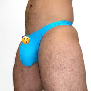 May include: A light blue thong swimsuit with a party emoji on the side. The swimsuit is made of a smooth, stretchy fabric. The background is plain white.