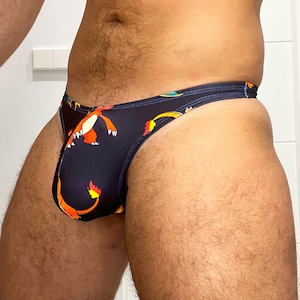 Puede incluir: Un slip de bikini negro con un patrón repetitivo de criaturas naranjas similares a dragones, con vientres amarillos y colas terminadas en llamas. La prenda cuenta con una cintura fina y aberturas de pierna altas con un diseño gráfico.