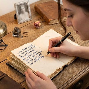 Op de afbeelding: Een persoon schrijft in een oud boek met een vulpen. Het open boek toont handgeschreven tekst. Een ingelijste foto, een inktpot, een bril en een gedroogde roos liggen op het houten bureau. De tekst in het boek luidt: "...mijn hart sloeg sneller toen onze ogen elkaar ontmoetten in de oude bibliotheek."