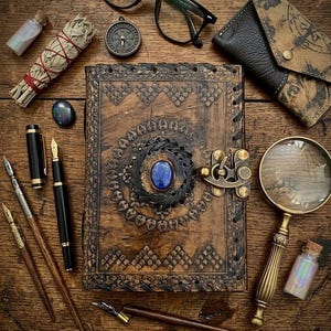 Op de afbeelding: Een bovenaanzicht van een houten tafel met diverse items. Een groot, ingewikkeld gesneden leren dagboek met een blauwe edelsteen staat centraal. Andere items zijn pennen, een vergrootglas, een bril, een kompas en een kleine tas.