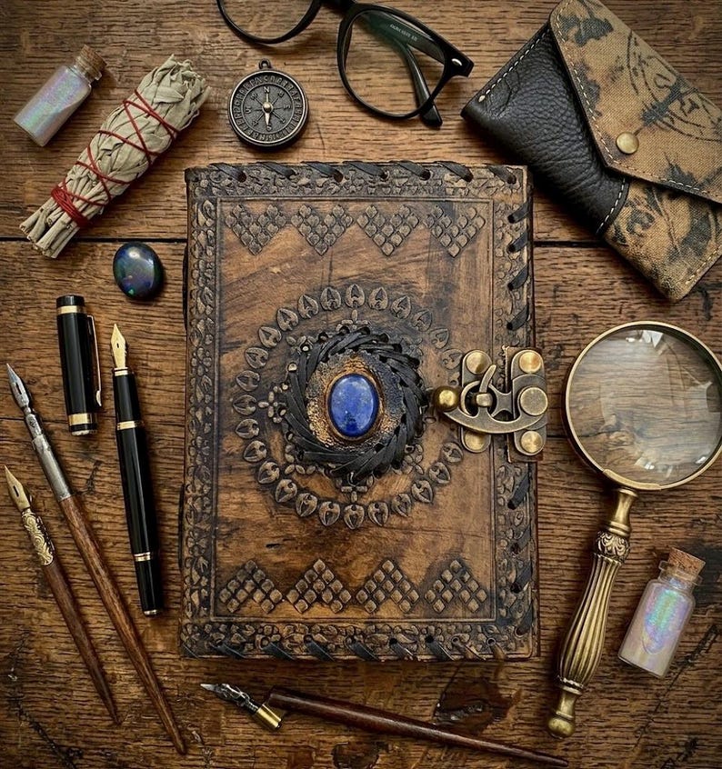 Op de afbeelding: Een bovenaanzicht van een houten tafel met diverse items. Een groot, ingewikkeld gesneden leren dagboek met een blauwe edelsteen staat centraal. Andere items zijn pennen, een vergrootglas, een bril, een kompas en een kleine tas.
