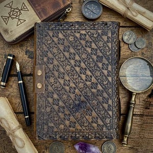 Op de afbeelding: Een vintage leren notitieboek met ingewikkelde bloempatronen, een leren riem en diverse antieke accessoires. Inclusief een kompas, vergrootglas, munten, pennen en een kleine portemonnee, allemaal gerangschikt op een houten oppervlak.