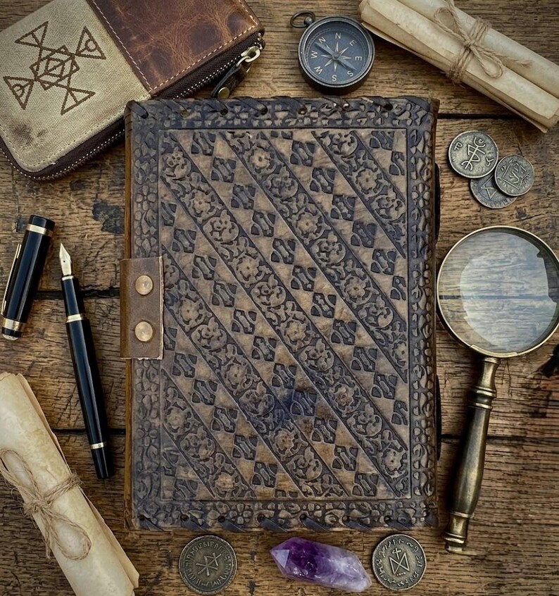 Op de afbeelding: Een vintage leren notitieboek met ingewikkelde bloempatronen, een leren riem en diverse antieke accessoires. Inclusief een kompas, vergrootglas, munten, pennen en een kleine portemonnee, allemaal gerangschikt op een houten oppervlak.
