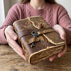 May include: A brown leather journal with a decorative floral design and a key closure. The journal is bound with a leather strap and features a key-shaped clasp. The pages have a vintage, aged appearance.