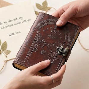 May include: A brown leather journal with an ornate embossed design and a metal clasp. The journal is being held by two hands, with a handwritten note in the background that reads, "To my dearest, my adventure awaits with you. With love."