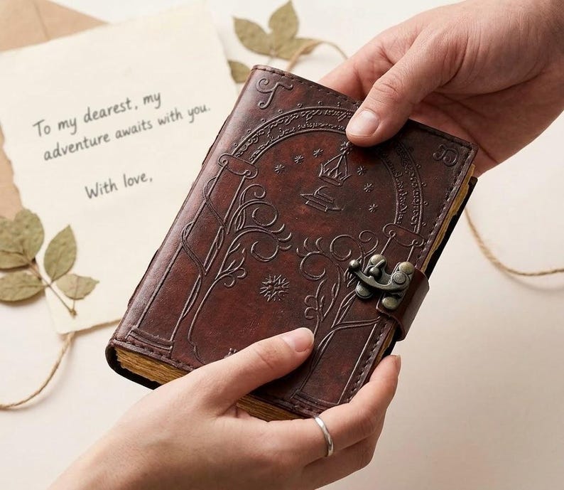 May include: A brown leather journal with an ornate embossed design and a metal clasp. The journal is being held by two hands, with a handwritten note in the background that reads, "To my dearest, my adventure awaits with you. With love."
