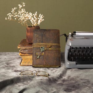May include: A leather-bound journal with a key clasp, a vintage typewriter, and a pair of round eyeglasses are arranged on a textured surface. A small wooden vase holds dried white flowers, and a stack of old books is visible.