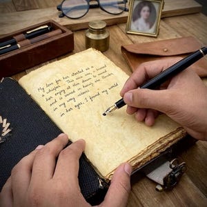 Op de afbeelding: Een zwarte vulpen schrijft op verouderd papier in een vintage dagboek. De tekst op de pagina luidt: "Mijn liefde voor haar begon als een fluistering... in haar ogen, vond ik mijn wereld." Andere items zijn een etui, een inktpot en een ingelijst portret.