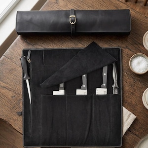 May include: A black leather knife roll with a brass buckle, open to show knives and a honing steel. The knives have black handles and silver blades. A matching leather roll is above. The knives are stored in a black fabric.