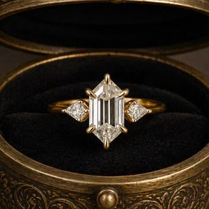 May include: A gold ring featuring a central elongated hexagonal-cut diamond flanked by two smaller kite-shaped diamonds, set in a yellow gold band and displayed inside a round, ornate, antique-style brass-toned metal box with black velvet lining.