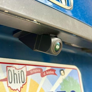 May include: A black backup camera mounted on a blue vehicle. The camera lens is visible, and the Ohio license plate is partially visible below, with the text "Ohio Birthplace of Aviation".