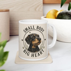 Könnte beinhalten: Weiße Keramik-Tasse mit einer Hundeillustration und dem Text "SMALL BODY BIG HEART". Der Hund ist schwarz und braun auf beigem Hintergrund. Die Tasse hat einen weißen Henkel und steht auf einem beigen Untersetzer. Die Tasse ist etwa 10 cm hoch.
