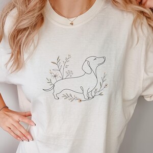 Könnte beinhalten: Cremefarbenes T-Shirt mit einer Strichzeichnung eines Dackels, umgeben von floralen Elementen. Der Hund ist in einem minimalistischen Stil dargestellt, und das Design ist auf dem Shirt zentriert. Die Gesamtästhetik ist schlicht und elegant.